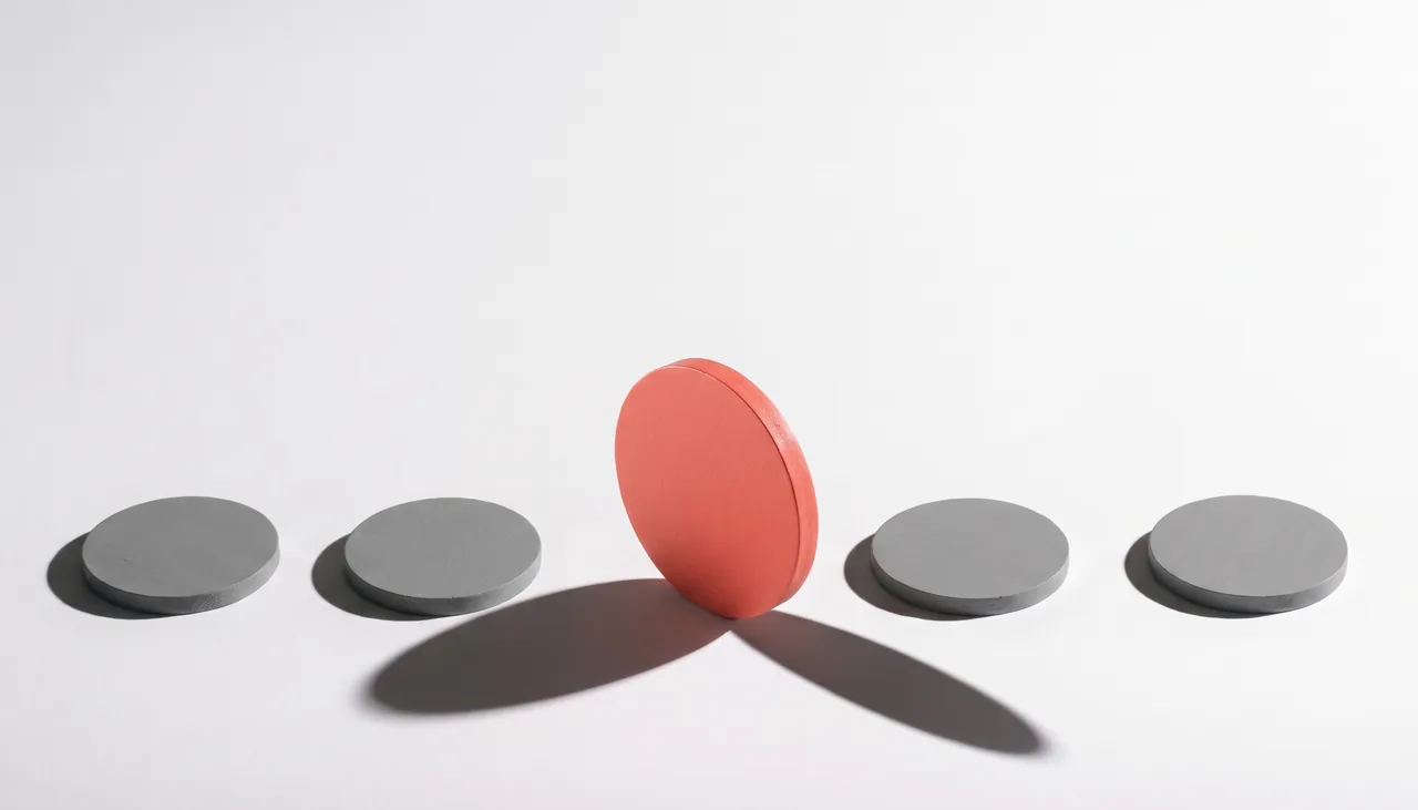 Conceptual still life: a row of identical flat grey discs on an off-white studio sweep, and one larger coral-red disc standing upright on its edge at the centre, casting a long hard shadow.